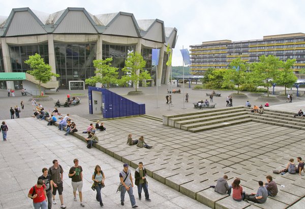 Foto: Ruhr-Universität Bochum The Audimax building features a concrete facade with distinctive angular roof sections and expansive glass windows. Several trees are visible in the foreground, along with flags indicating an academic setting.
