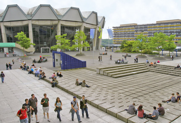 Foto: Ruhr-Universität Bochum The Audimax building features a concrete facade with distinctive angular roof sections and expansive glass windows. Several trees are visible in the foreground, along with flags indicating an academic setting.