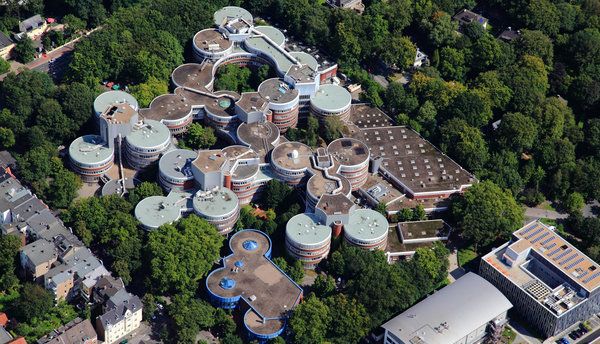 Photo: University Duisburg-Essen, © https://www.flickr.com/photos/57915984@N04/23818975739/ Aerial perspective of a complex of interconnected circular buildings, featuring green rooftops and surrounded by lush trees. The structures form a unique architectural pattern, integrating seamlessly with the natural environment.