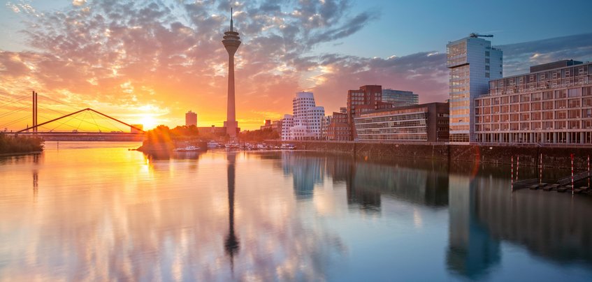 Foto: Max-Planck-Institut für Nachhaltige Materialien GmbH A colorful sunset casts warm hues over Düsseldorf's skyline, with the Rheinturm tower and modern architecture mirrored in the calm Rhine River water.