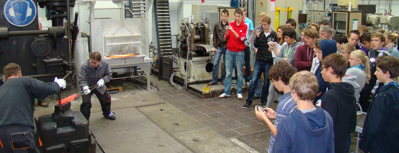 Mitarbeiter des MPIE führen den Schmiedehammer vor. Die deutschen und niederländischen Schüler zeigen sich beeindruckt. Im Hintergrund ist der Ofen erkennbar, in welchem das Metall auf über 1000 °C erhitzt wird.