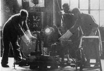 Employees of the institute's department of Metallurgy at the high frequency casting, 1920. Three individuals in protective gear work on machinery in a workshop, illuminated by a bright light.