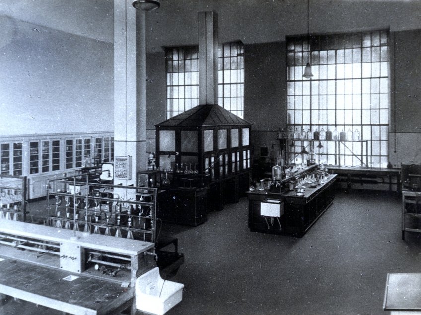 A view of the chemical laboratory in the old institute building. Large laboratory with a central glass enclosure, surrounded by tables with scientific apparatus.