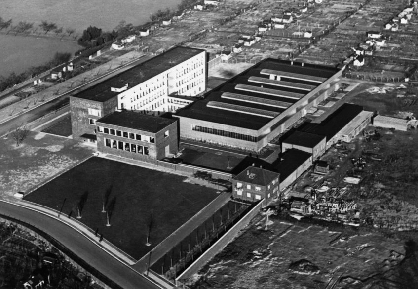 This aerial view shows the main building, the hall construction and the auxiliary buildings after completion in the autumn of 1935. Aerial view of a large industrial complex with multiple buildings and surrounding roads.