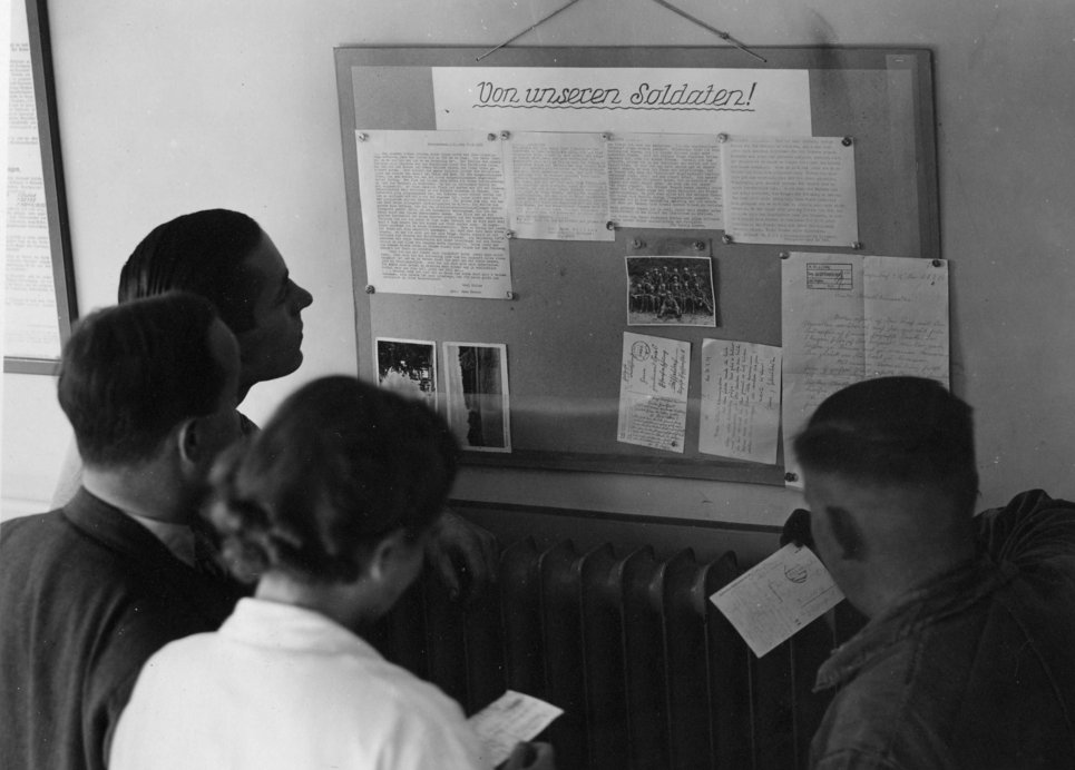 Mitarbeiter und Mitarbeiterinnen studieren die „Feldpostbrief-Tafel“ des Instituts, an der die Briefe und Karten der zum Krieg eingezogenen Arbeitskollegen ausgehängt wurden.
