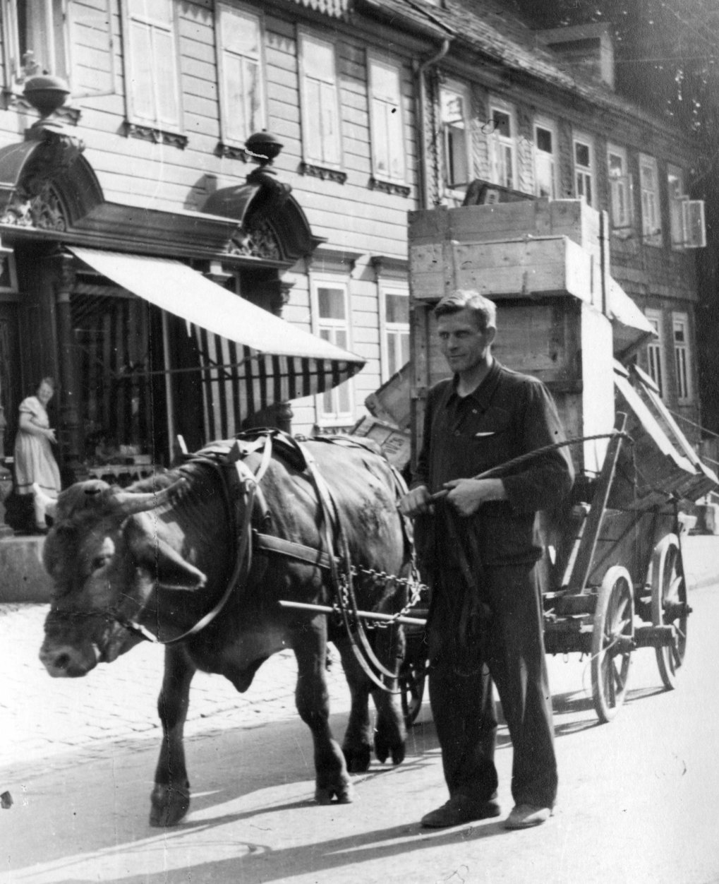 A man guides an ox pulling a cart with stacked crates on a cobblestone street in front of old buildings.