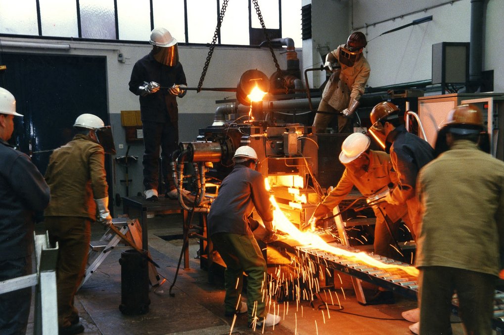 Workers in protective gear operate machinery with glowing metal and sparks.