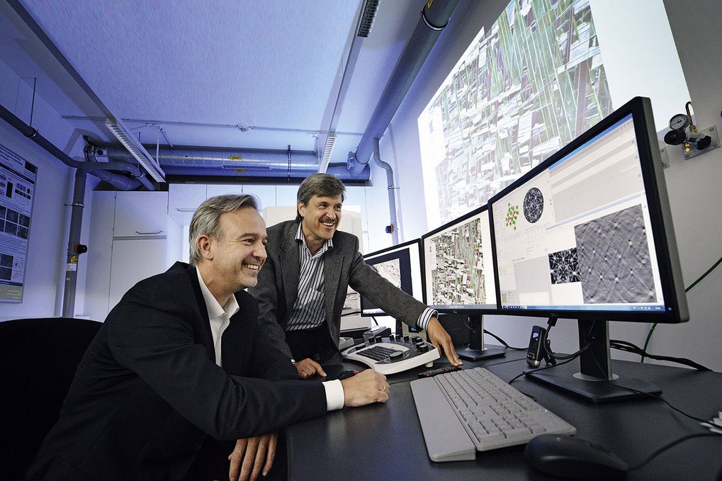 Two people in a modern office examining complex data on three computer monitors and a large projection.