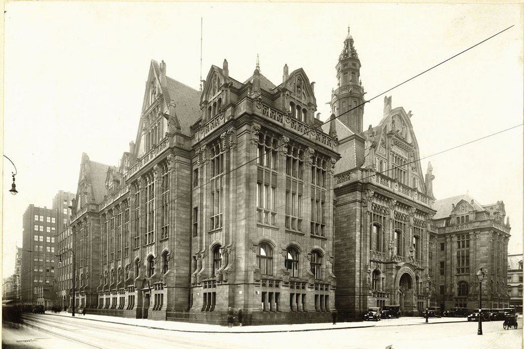 A grand historical building with multiple gables, towers, and detailed stonework, situated on a busy street corner.