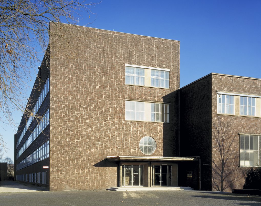 A modernist brick building with symmetrical large windows, a prominent circular entrance feature, and a stark shadow cast on the facade under a clear sky.