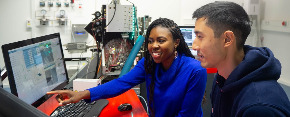 In a laboratory, two persons are sitting in front of computer screens, one is pointing at a screen. Scientific devices are seen in the background