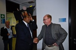 Person dressed in a suit is shaking hands with another, wearing a large black hat adorned with various small items, in a room with visible office elements like maps and signage.