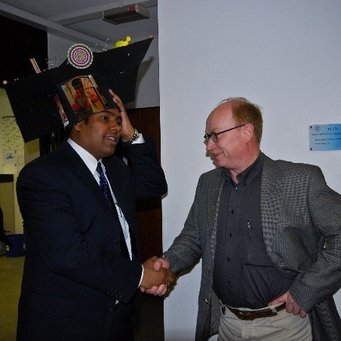 PhD Defense on 12th June 2008 Person dressed in a suit is shaking hands with another, wearing a large black hat adorned with various small items, in a room with visible office elements like maps and signage.
