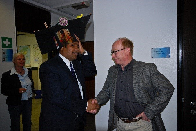 Person dressed in a suit is shaking hands with another, wearing a large black hat adorned with various small items, in a room with visible office elements like maps and signage.