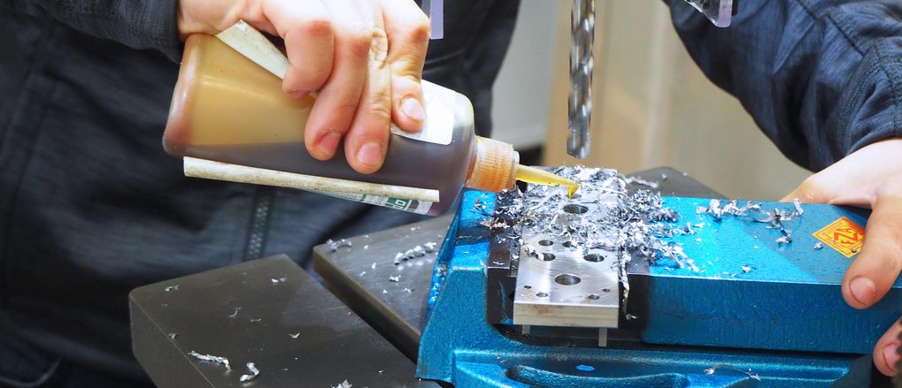 Industriemechaniker*inFachrichtung Feingerätebau(m/w/div) Metall wird mit einer Bohrmaschine bearbeitet, während Schmiermittel hinzugefügt wird.