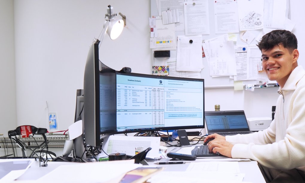 Kauffrau/-mann für Büromanagement(m/w/div) Person am Schreibtisch mit Computer, umgeben von Dokumenten und zwei Bildschirmen.