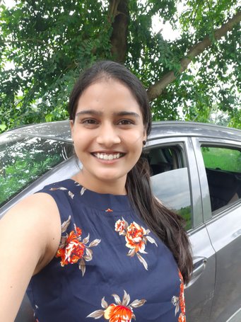 A woman in a navy blue, sleeveless floral dress stands beside a gray car with an open door, surrounded by vibrant green foliage and trees.