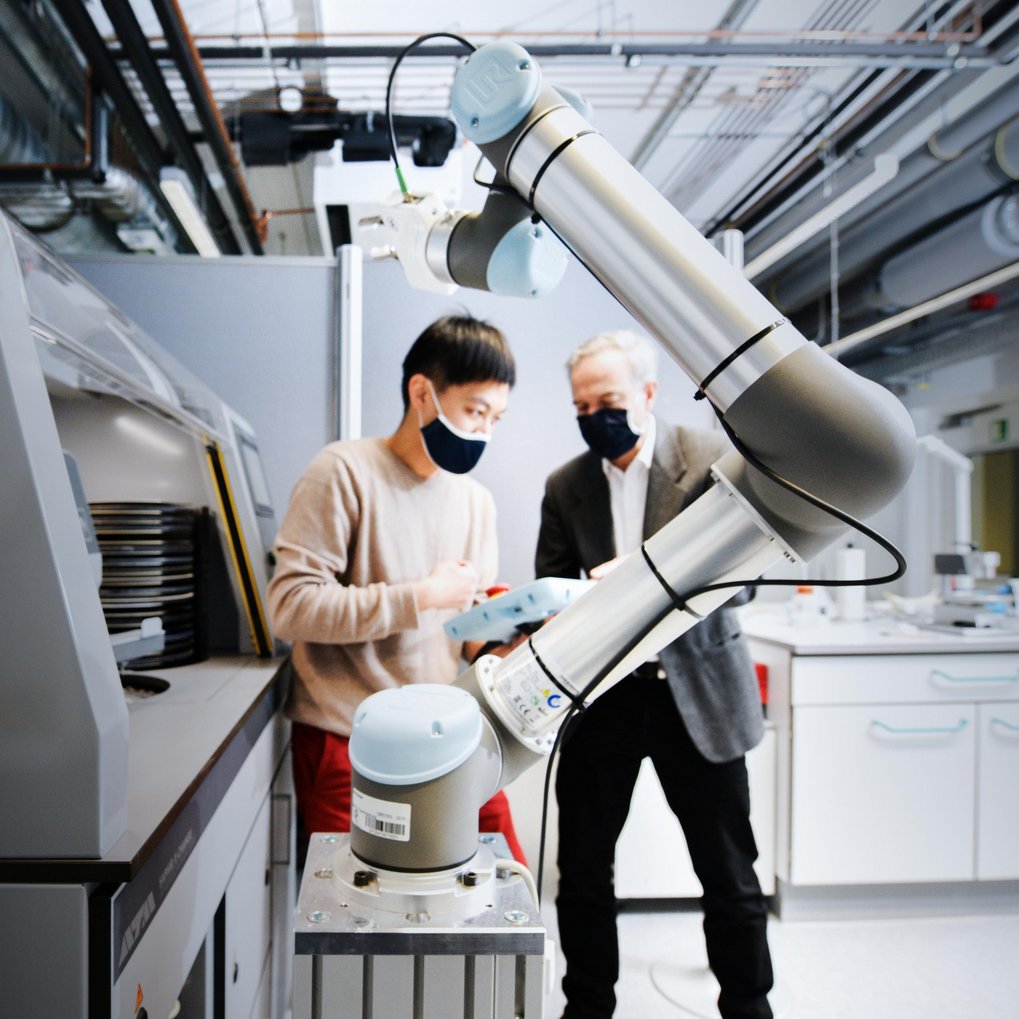 A robotic arm is seen in the foreground. Two scientists are looking at a tablet in the background.