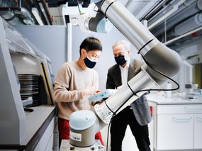 A robotic arm is seen in the foreground. Two scientists are looking at a tablet in the background.