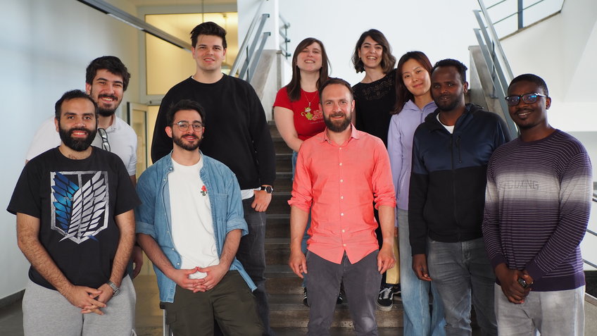 © Max-Planck-Institut für Nachhaltige Materialien GmbH A diverse group of ten people casually positioned on indoor stairs, featuring varied casual clothing styles and neutral expressions.