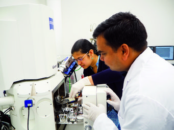 Two scientists, wearing lab coats and gloves, manipulate a sample within an advanced Zeiss Gemini electron microscope in a laboratory setting, focusing on precision and detail.