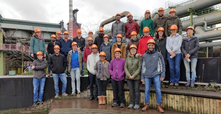 © Max-Planck-Institut für Nachhaltige Materialien GmbH Large group wearing orange safety helmets, standing in front of industrial structures with visible pipes and buildings.