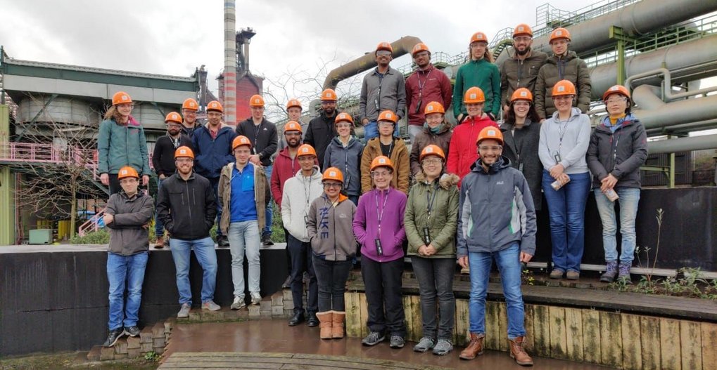 A group of people wearing orange hard hats, standing in front of industrial pipes.