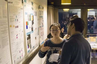 An indoor scene featuring two individuals in focused discussion, analyzing various scientific posters with charts and graphs on a wall.