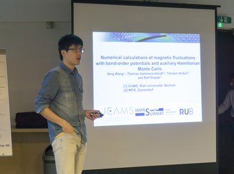 A researcher stands beside a projected slide detailing numerical calculations of magnetic fluctuations with bond-order potentials and auxiliary Hamiltonian Monte Carlo methods. The slide lists contributors from ICAMS, Ruhr Universität, and MPIE Düsseldorf.