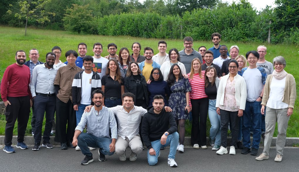 Diverse group of men and women posing together on a paved road, with a background of lush green grass and trees, suggesting a casual outdoor setting.