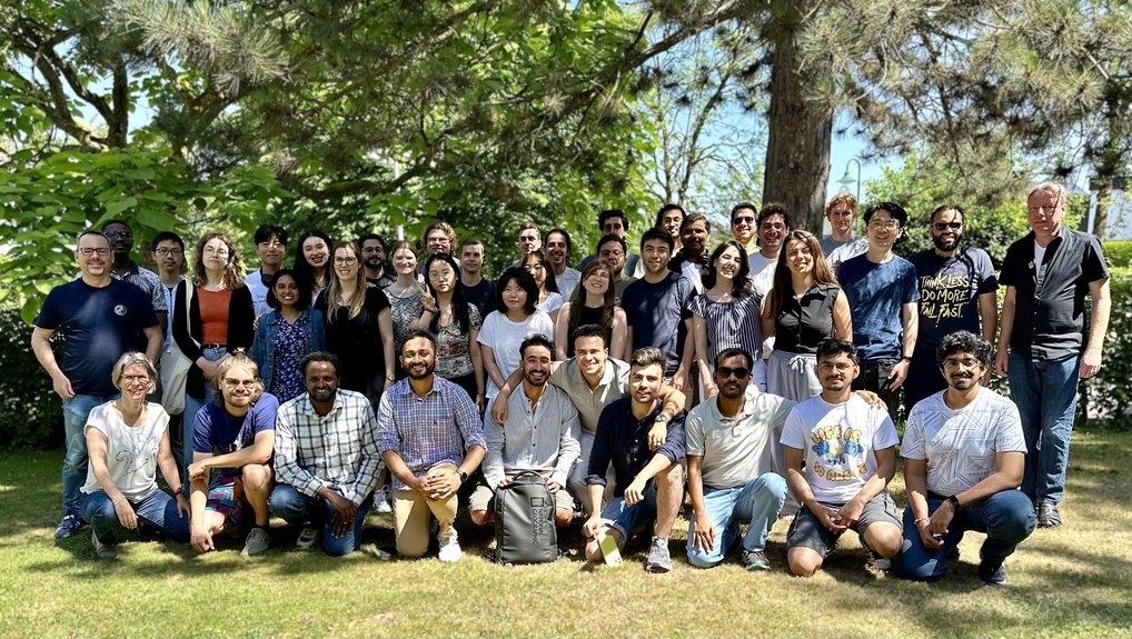 A mixed group of individuals is gathered outdoors on a grassy area, with several trees providing shade, suggesting a casual group photo.
