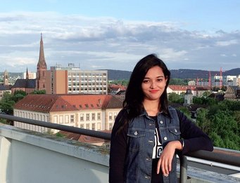 Individual standing on a rooftop terrace overlooking a cityscape with a prominent church steeple and various modern buildings, set against a backdrop of a blue sky and scattered clouds.