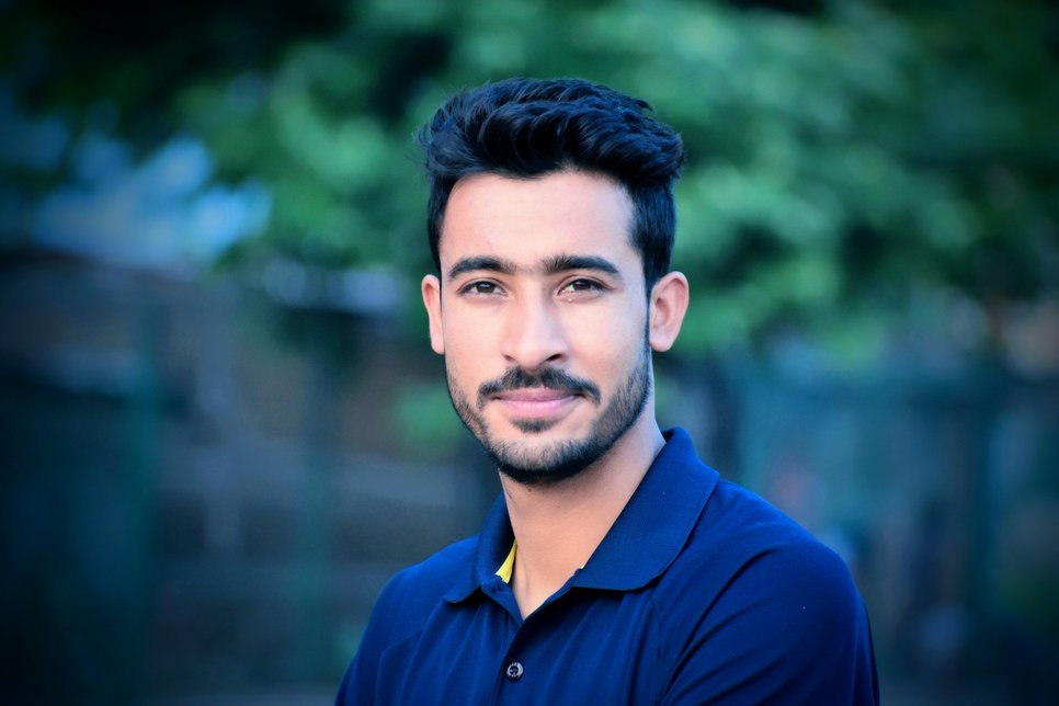 An individual in a blue polo shirt stands with arms crossed, against a blurred backdrop of green foliage and fencing.