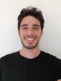 Individual with dark, curly hair wearing a simple black shirt, positioned against a plain, light-colored wall, showing upper torso.
