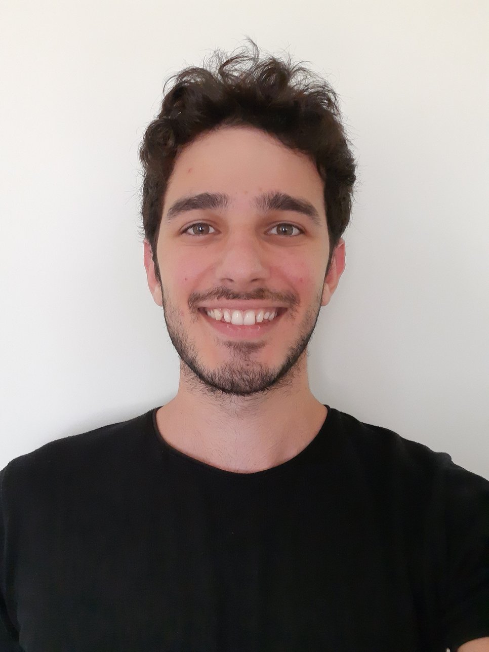 Individual with dark, curly hair wearing a simple black shirt, positioned against a plain, light-colored wall, showing upper torso.