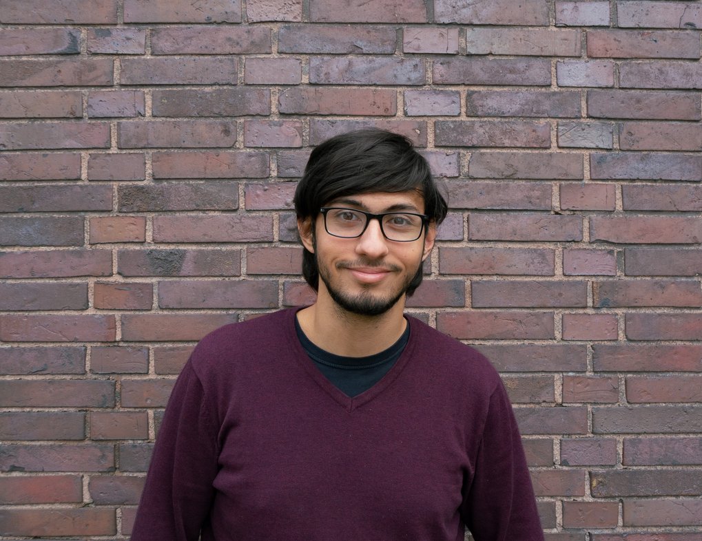 Person with dark hair in a burgundy sweater, situated in front of a textured brick wall. The bricks vary in shades of brown and gray.