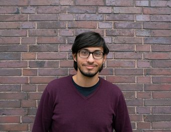 Person with dark hair in a burgundy sweater, situated in front of a textured brick wall. The bricks vary in shades of brown and gray.