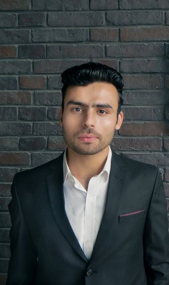 Person dressed in a formal black suit and white shirt, positioned against a textured brick wall backdrop.