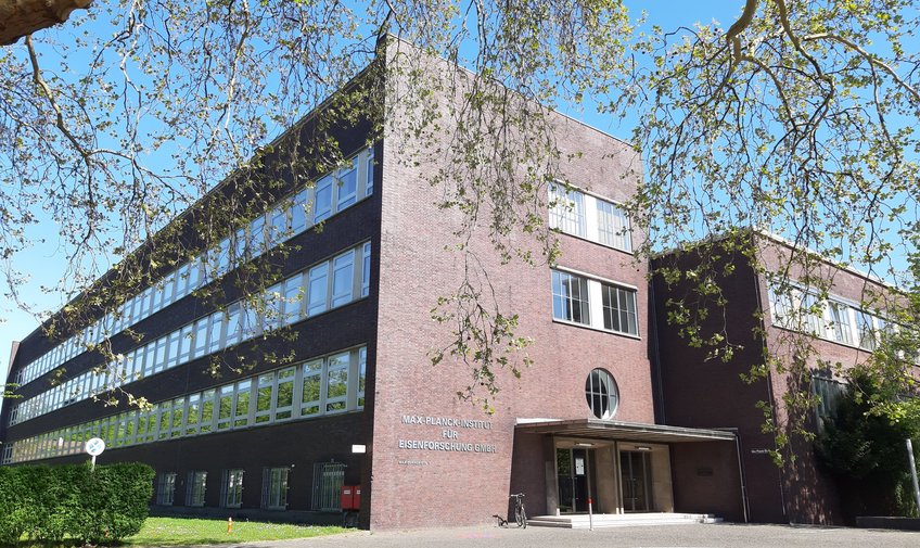Max-Planck-Institut für Nachhaltige Materialien, Foto Navyanth Kusampudi A modernist brick building with symmetrical large windows, a prominent circular entrance feature, and a stark shadow cast on the facade under a clear sky.