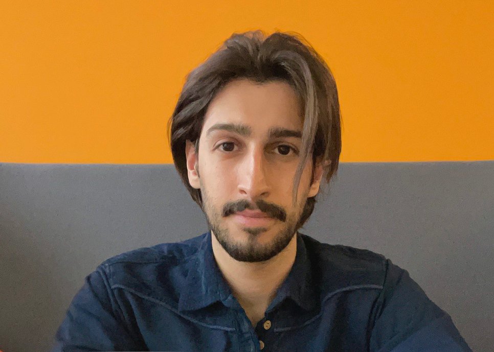 A person with a dark blue shirt in front of an orange wall while leaning against a gray surface. The hair is medium length and dark brown.