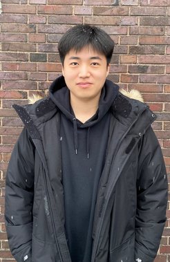 A person is standing in front of a brick wall and is wearing a black winter jacket with a hood on which snowflakes can be seen.
