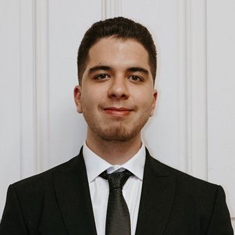 A man dressed in a black suit and tie with a white shirt, standing against a white paneled wall, exuding a professional appearance.