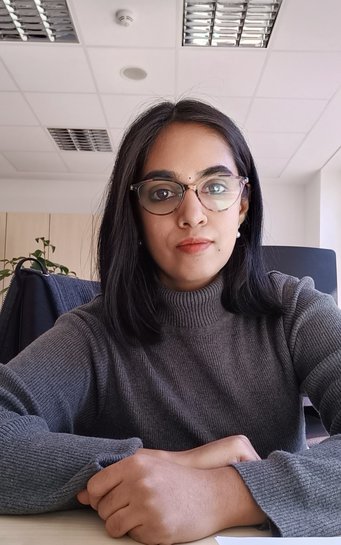 Person wearing a gray sweater sits at a desk in an office. The ceiling features recessed lighting panels, and a plant is visible in the background.