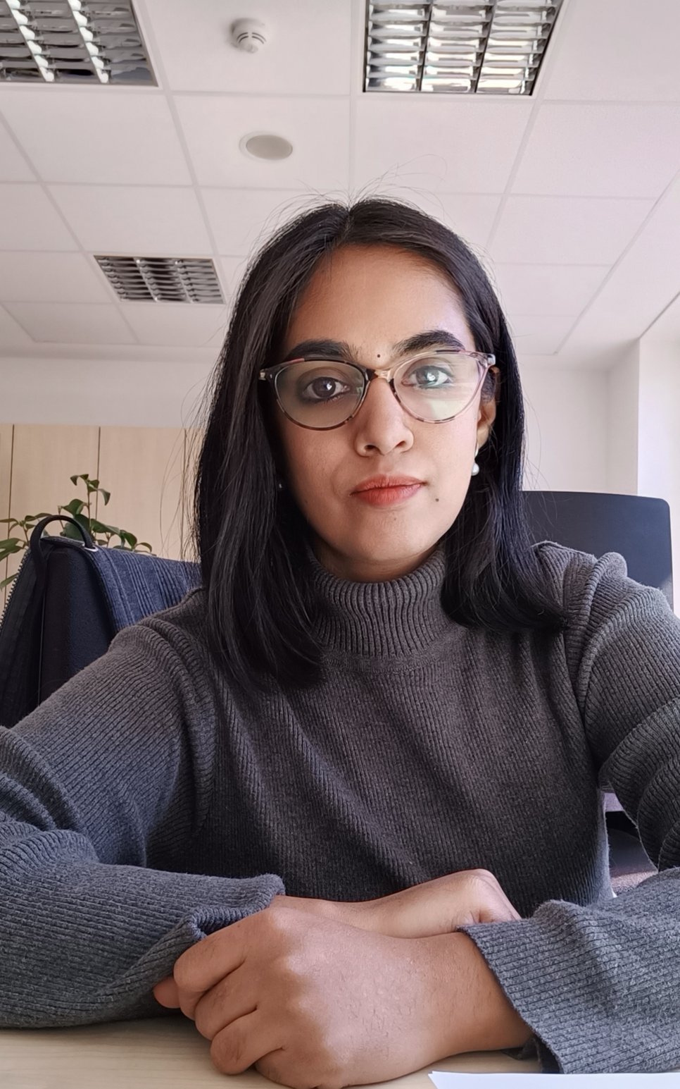 Person wearing a gray sweater sits at a desk in an office. The ceiling features recessed lighting panels, and a plant is visible in the background.
