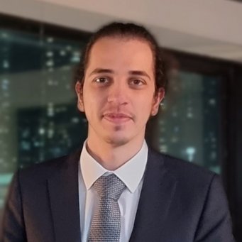 A person wearing a dark suit and textured tie is positioned in an interior space, featuring a large window that offers a glimpse of a cityscape with numerous lit windows.