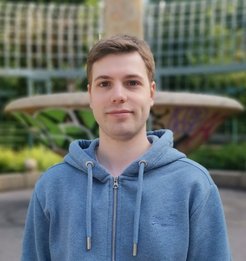 A person wearing a blue zip-up hoodie with drawstrings stands outside near a decorative fountain, face blurred.