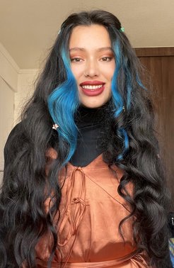 Black hair with blue highlights styled in waves, adorned with small clips, paired with an orange dress featuring intricate ribbon ties at the front.