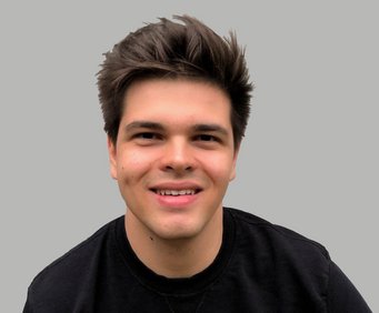 The subject has dark hair styled upwards and wears a black shirt. The background is a neutral gray, providing contrast to the attire and hair.