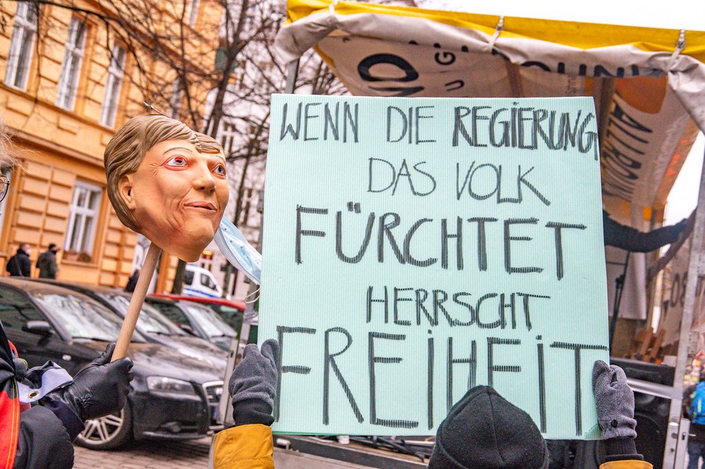 Demonstration mit kleiner Tribüne, davor ein Schild mit der Aufschrift "Wenn die Regierung das Volk fürchtet, herrscht Freiheit" und ein Stab, auf dem eine Gummimaske mit den Gesichtszügen von Angela Merkel hochgehalten wird