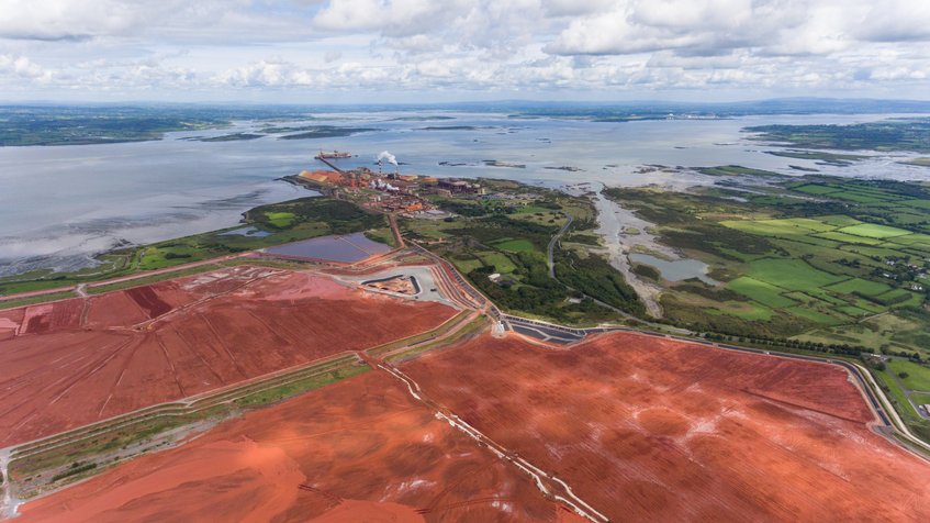 The corrosive and toxic red mud produced during aluminium production is currently disposed of in large landfills, such as here at the Aughinish aluminium smelter in Ireland. A team from the Max-Planck-Institut für Eisenforschung has developed an economical process to use it to produce climate-neutral iron for the steel industry.  In the foreground, a section of the extensive area of a landfill site with rust-red mud, in the background a much smaller aluminium plant. The plant and landfill are located on a gulf, which can be seen in the upper half of the picture. Green meadows can be seen on the right.